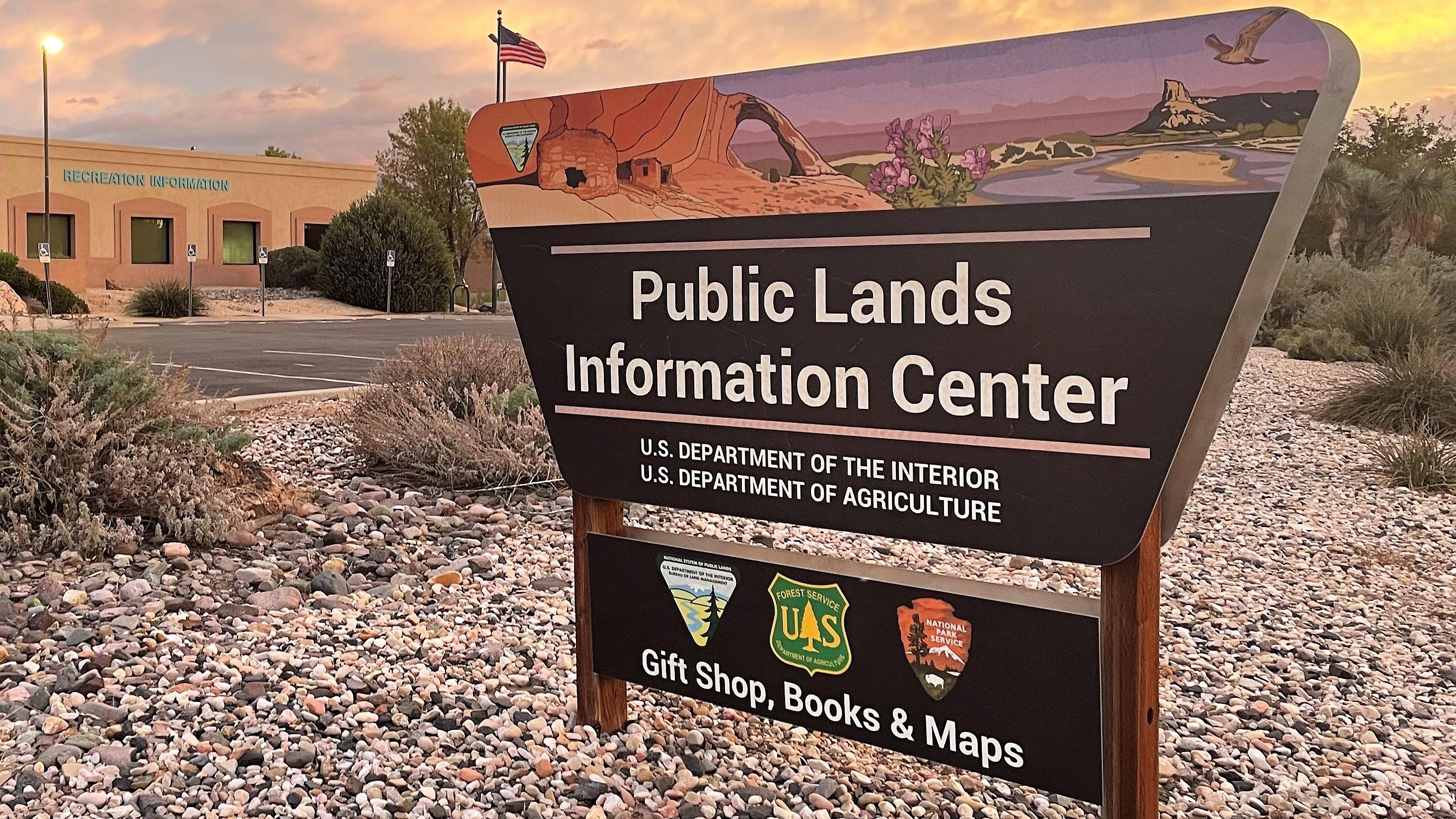 A sign stands in front of an office building and parking lot with an American flag flying overhead.