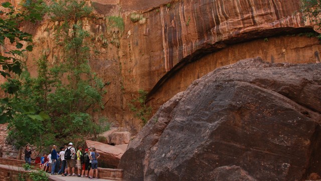 Group of visitors are standing on a paved path looking at a large, red sandstone arch on a wall.