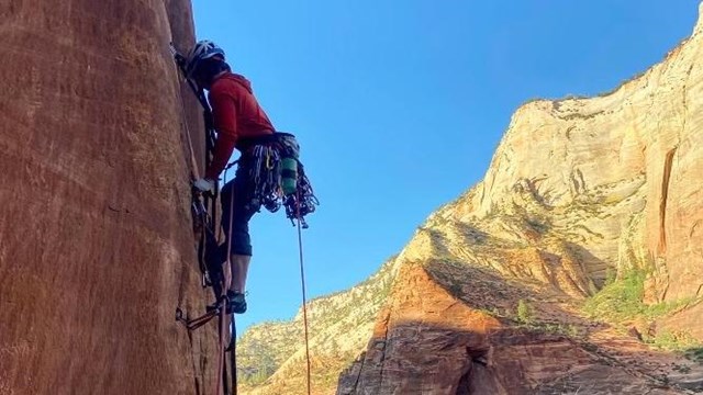 A climber scales a vertical wall high above the canyon floor.