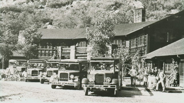 A black-and-white photo of old cars parked in front of a lodge. Sandstone cliffs stand in the back.