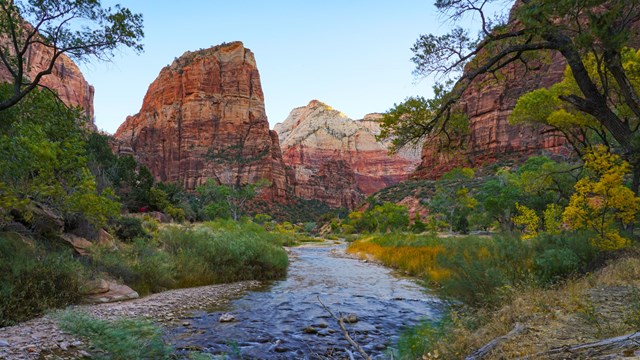 A tall sandstone peak stands above a blue, flowing river in a deep canyon. Trees line the river.