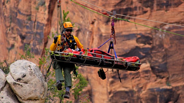A man in a yellow, long-sleeve shirt and orange helmet descends a rope with a metal stretcher.
