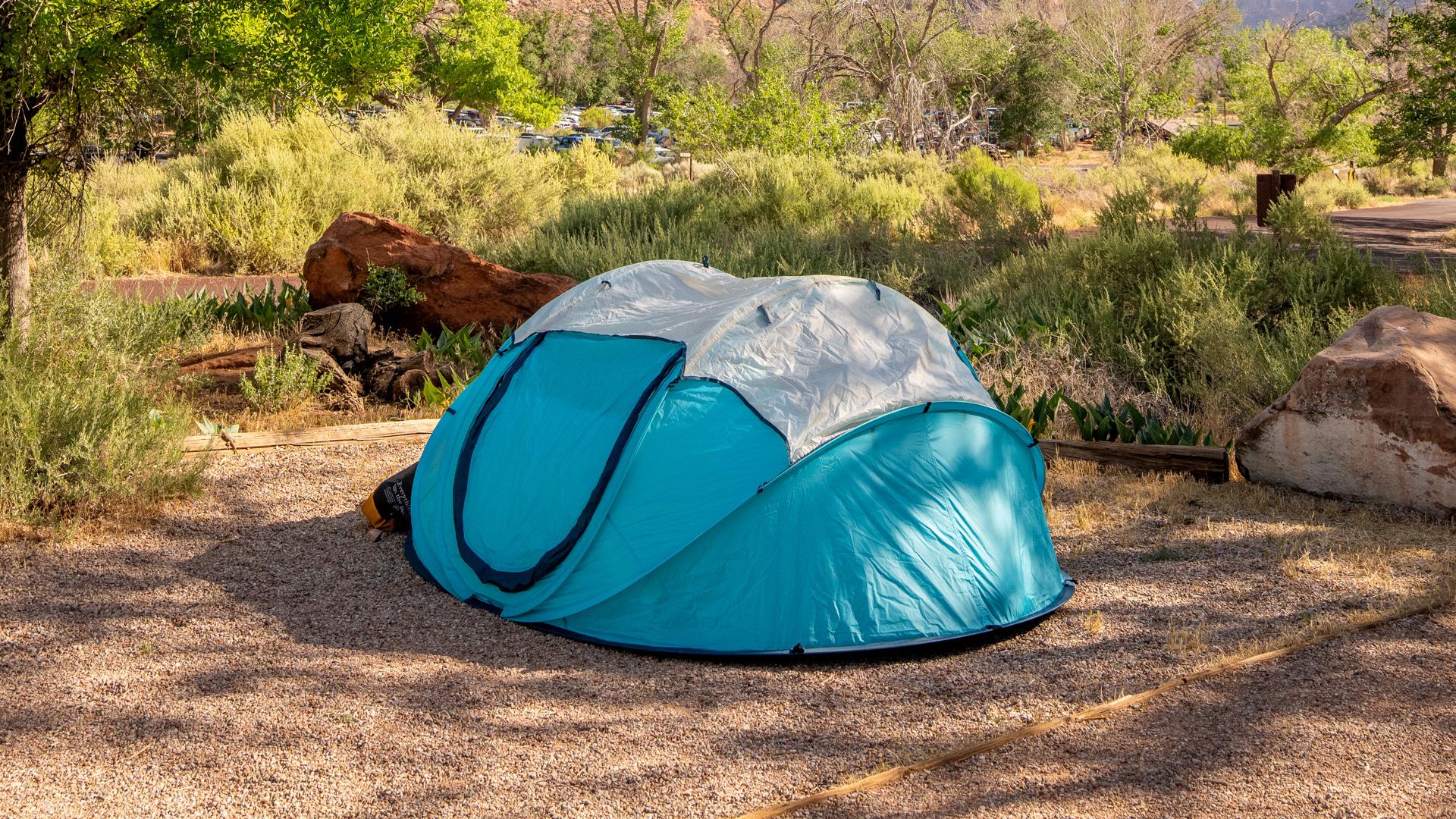 A bright blue tent sits in a campsite. Small trees and huge sandstone cliffs are visible.