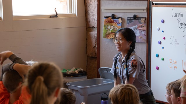 A park ranger with glasses kneels and talks to a group of children.