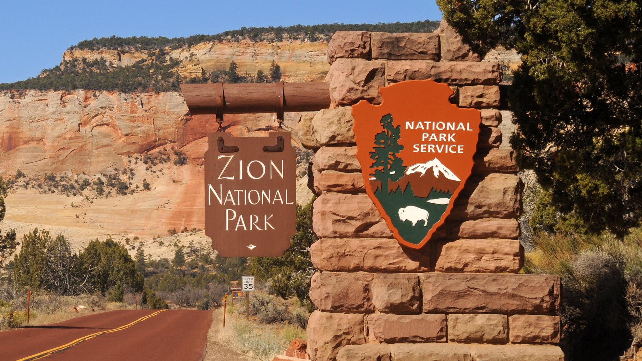 The Zion entrance sign, with a red road and red sandstone cliffs in the background. 