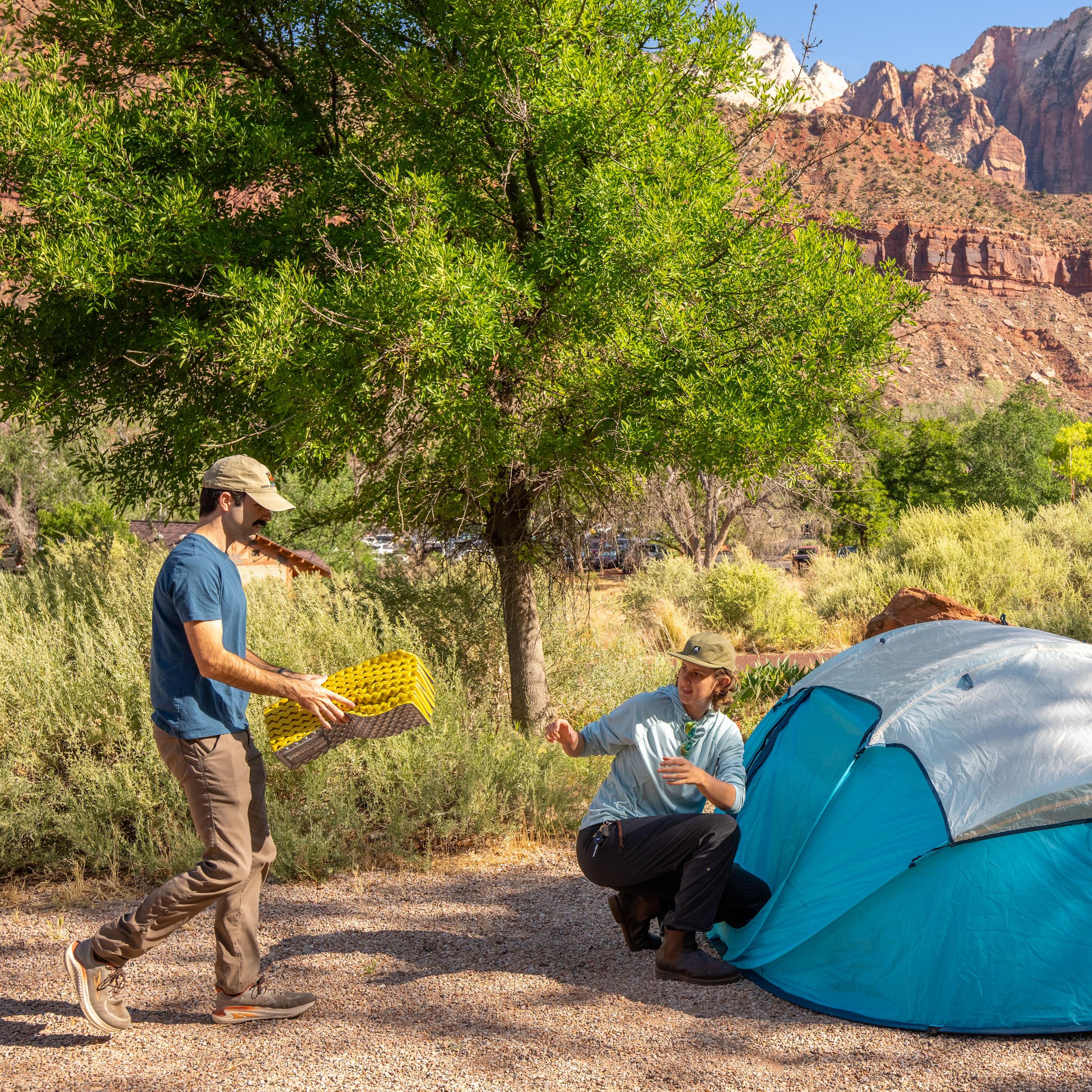 Two people set up camp next to a bright blue tent. Huge sandstone cliffs stand in the background.