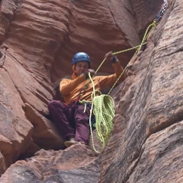 A rock climber wearing a blue helmet gathers up a climbing rope while standing on a cliff edge.