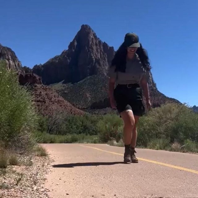 A ranger walks quickly down a cement path, sandstone cliffs in the background.