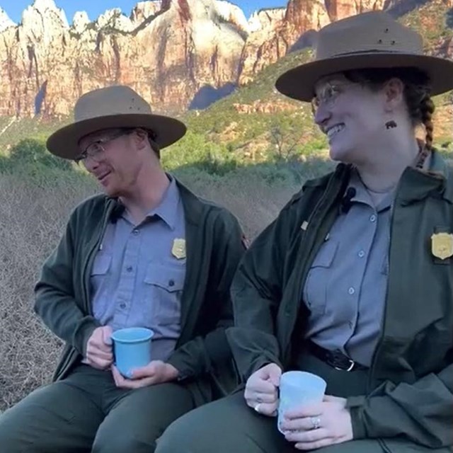 Two smiling rangers sit at shuttle stop while holding coffee mugs.