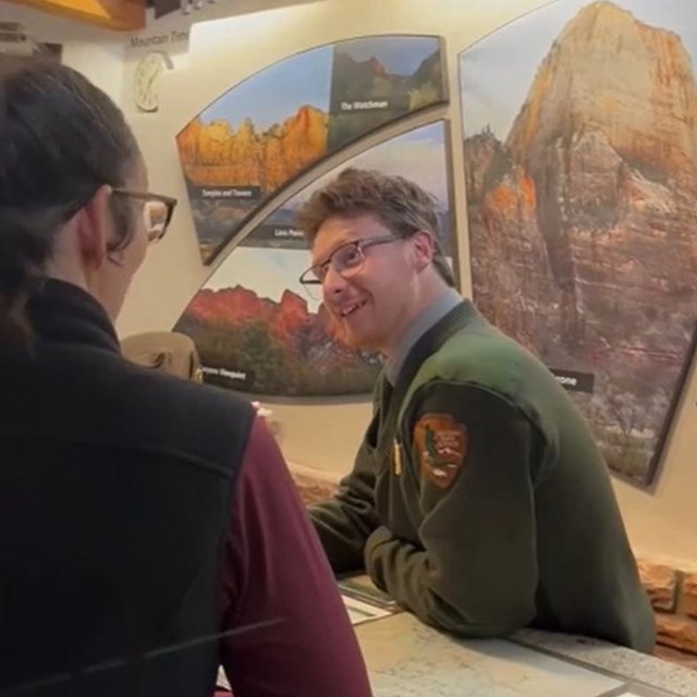 A ranger in green and gray speaks to a visitor from behind the visitor center information desk.