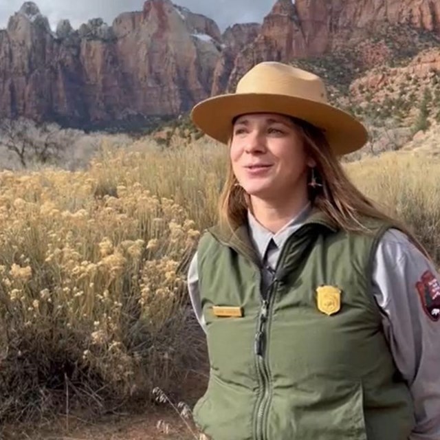 A ranger with long blonde hair speaks in front of a desert landscape of sandstone cliffs and bushes.