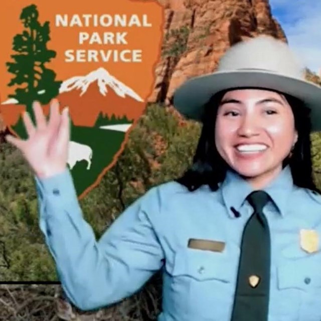 A ranger smiles and waves in front of a backdrop of a National Park Service arrowhead logo.