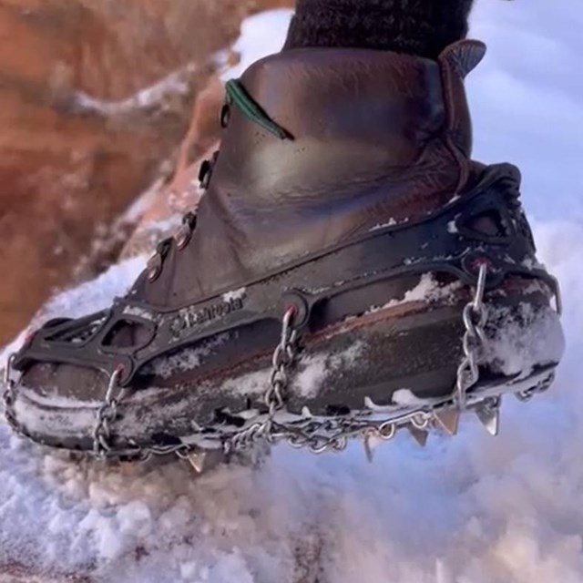 A foot, wearing a brown boot with metal micro-spikes for traction, steps up a snowy rock.