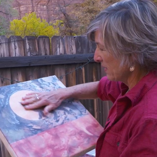 An artist smears her hands across a canvased collage of a fox, a full, moon and abstract landscape.