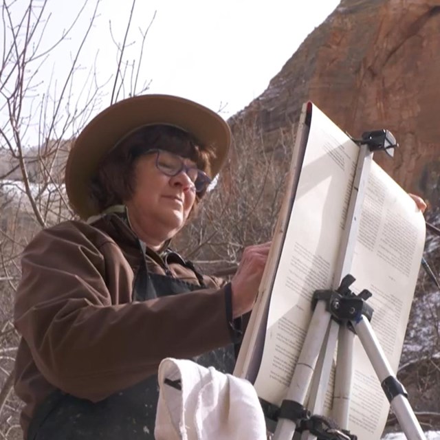 An artist stands and paints outside in a landscape of leafless trees and snow-capped cliffs.