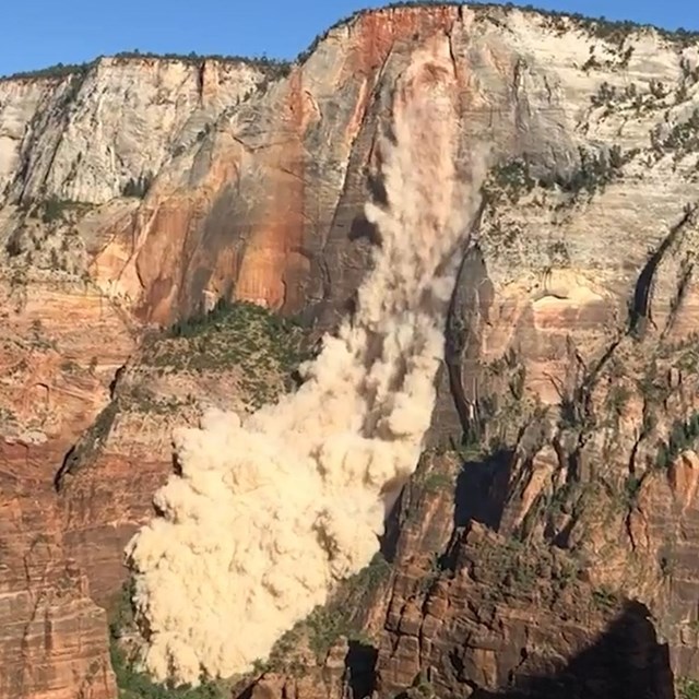 A massive, tan cloud of dust rolls down a red sandstone cliff, engulfing the green plants below.