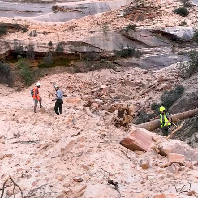 Three rangers in neon protective equipment scramble on top of a large pile of rock, sand, and dust.