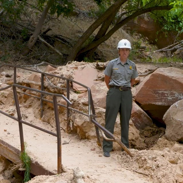 A ranger in a hard hat stands on a walkway that's railings are destroyed by large boulders.