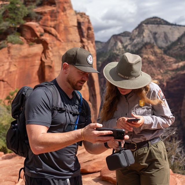 A ranger looks at a man's phone with a canyon and orange sandstone in the background.