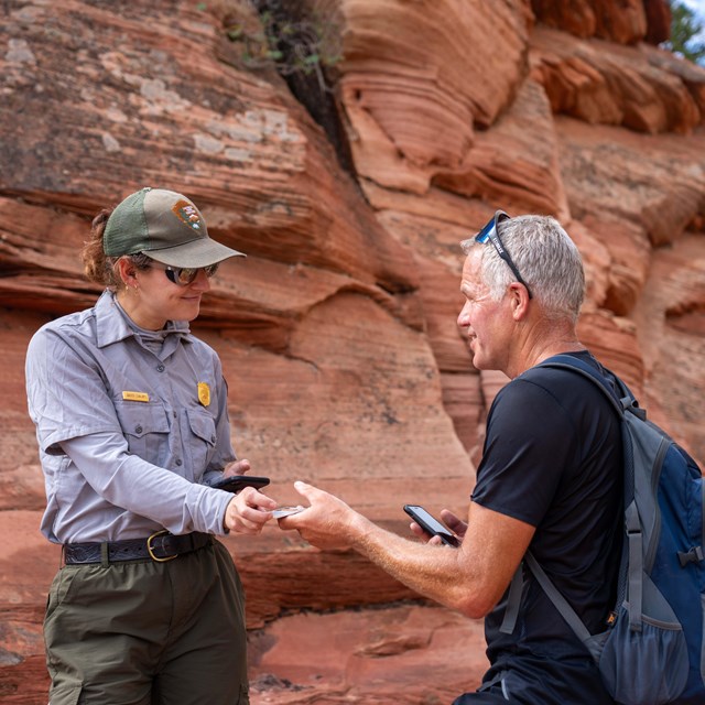 A ranger smiles and hands a man an ID next to orange sandstone cliffs