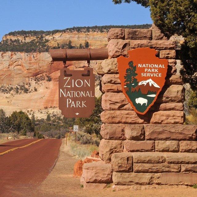 The Zion entrance sign, with a red road and red sandstone cliffs in the background. 