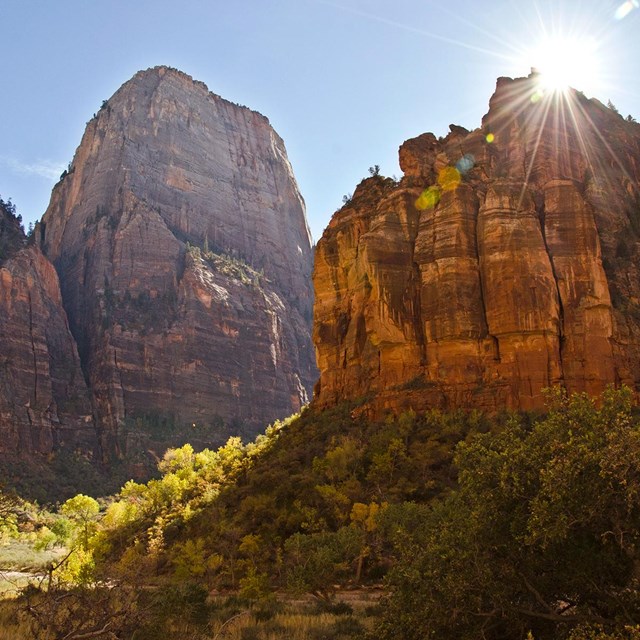 Sunshine peaking out behind a red sandstone cliff. A tall mountain is in the background.