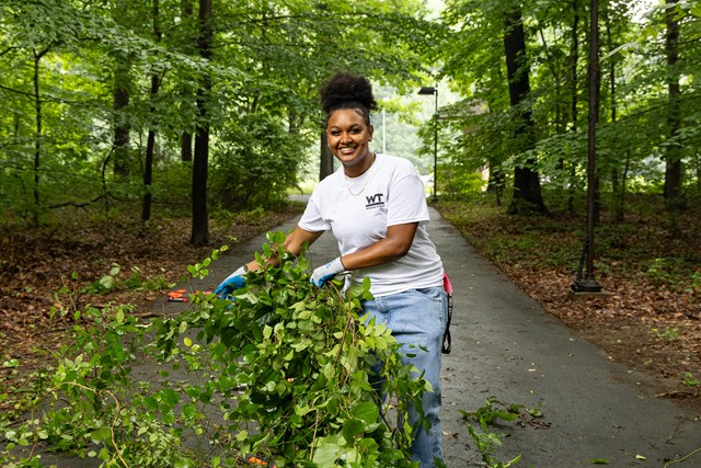 A smiling young woman holds a large clump of leafy plant material on a woodland trail.