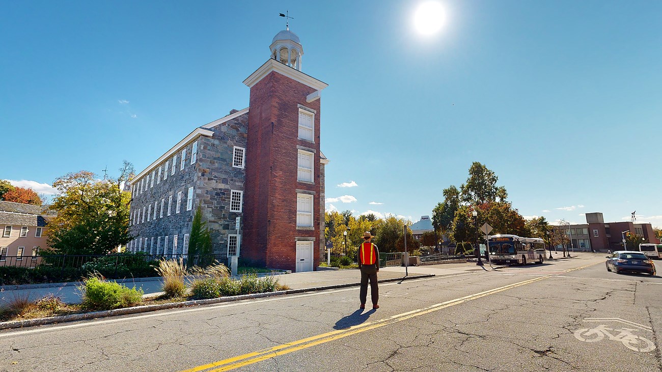 Virtual tour screenshot of a ranger standing in the middle of the road, and a top-to-bottom view of Wilkinson Mill just beyond. The mill features a red brick entrance portion topped with a little bell tower. The rest of the three-story building is stone.