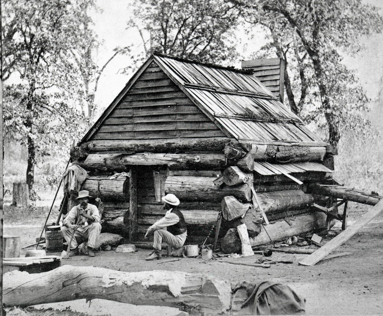 Two people sit in front of a log cabin. 