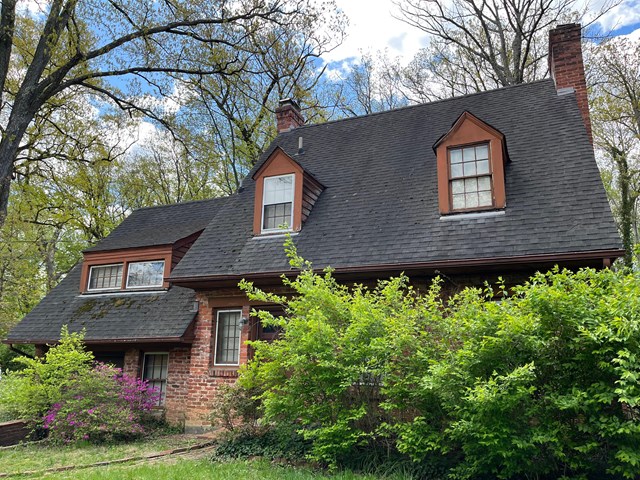a front view of a two-story residential building with a long, slanted roof with shingles and dormers. Due to the angle, it is mostly obscured by bushes in front of the house.