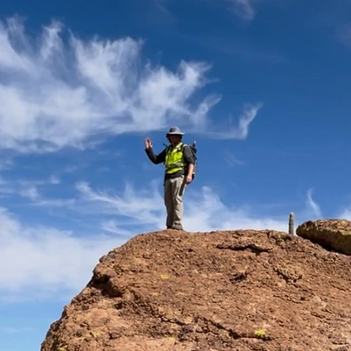 A man in field clothes poses on top of a large boulder with a waving hand.