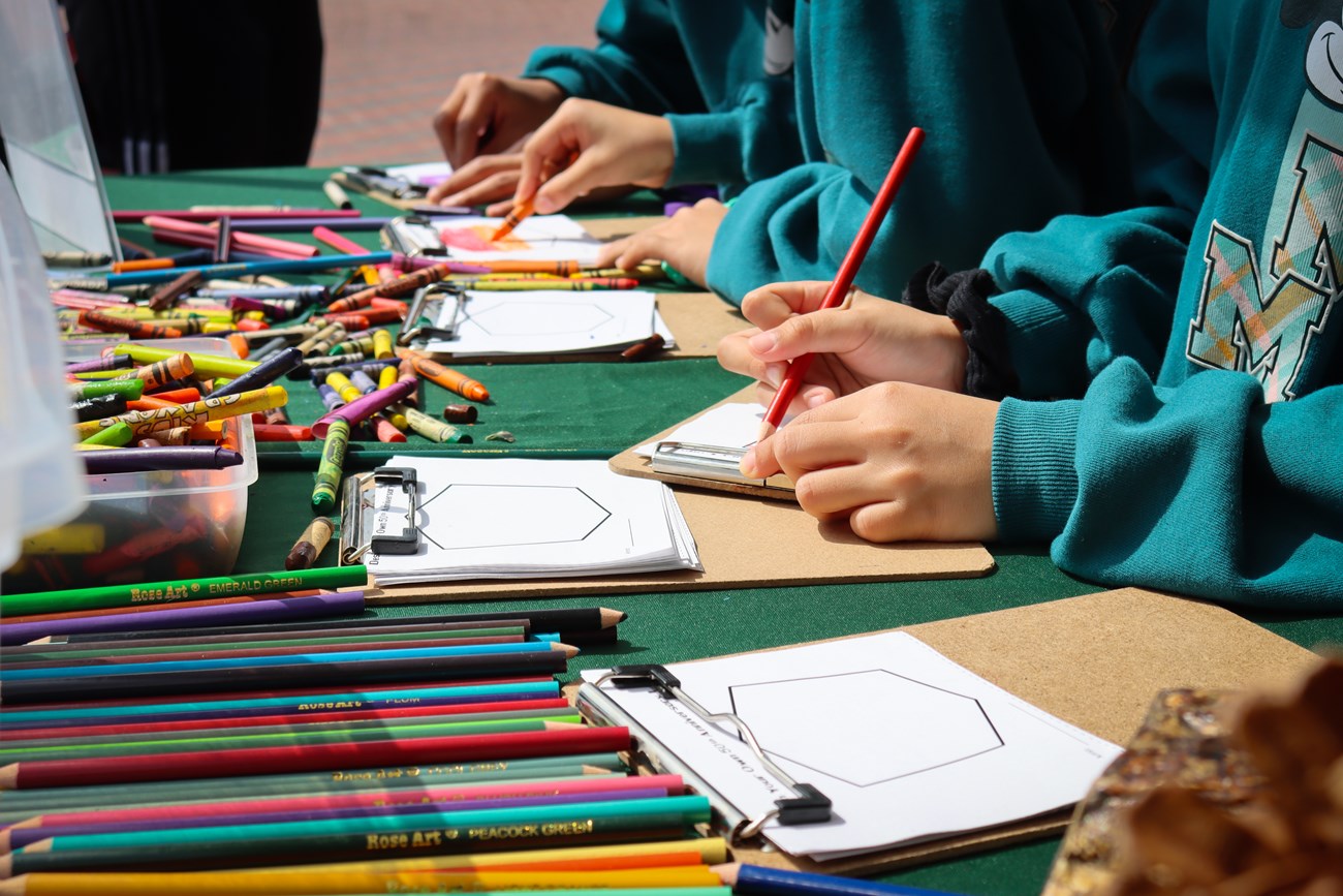 children using colored pencils and crayons to draw on small pieces of paper on a clipboard.