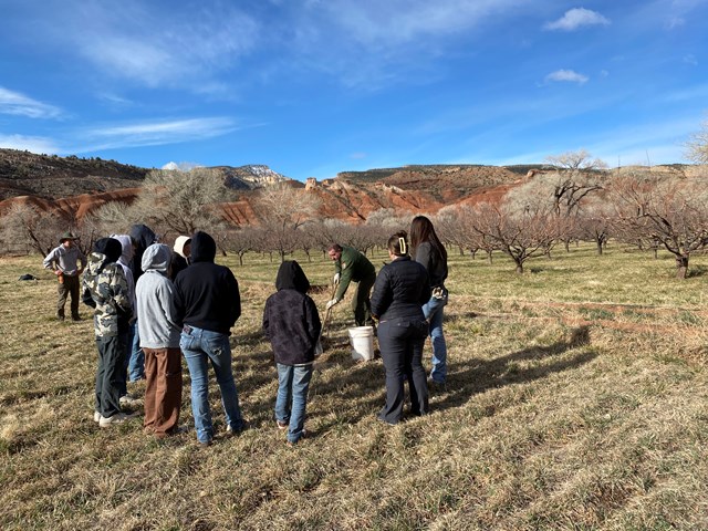 Students stand in a semicircle around a man digging in an open field.