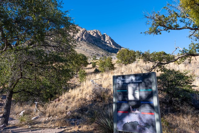 trailhead sign with rocky dry terrain, trees, and mountain