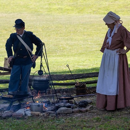 Man dressed in civil war uniform with woman dressed in 19th century dress, standing around a fire wi
