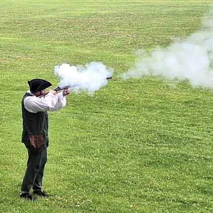 Man dressed in 18th century clothing, firing a musket in a field with smoke coming out of the gun. 