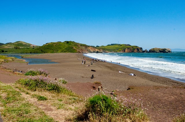 People sit in the sand on a beach as waves roll in.
