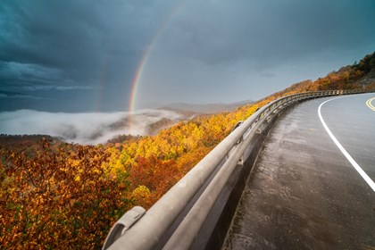 A roadway overlooking mountains of changing fall leaves with a rainbow casted over a cloudy sky w