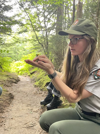 A ranger holds a bird on her palm about to release it, while sitting beside a trail.