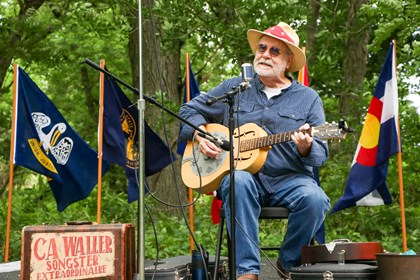 Man in blue shirt and jeans sits on stage playing the guitar. State flags fly behind him and a sign in front reads, 