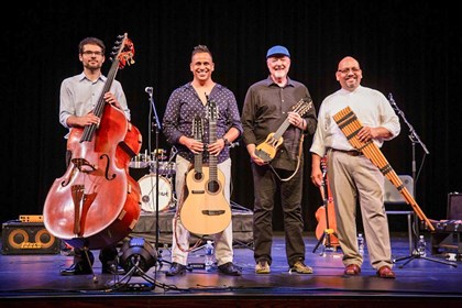 Four smiling men stand on a stage looking out. The three on the left hold diverse stringed instruments. The one on the right holds a woodwind.