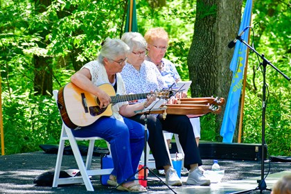 Three ladies play stringed instruments on stage. The one on the left plays the guitar; the two on the right play dulcimers.