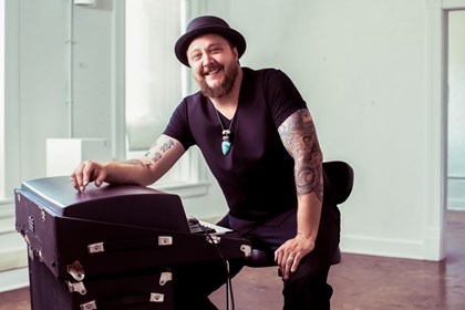 A smiling, seated, bearded young man in black with arm tattoos looks into the camera from behind a piano.