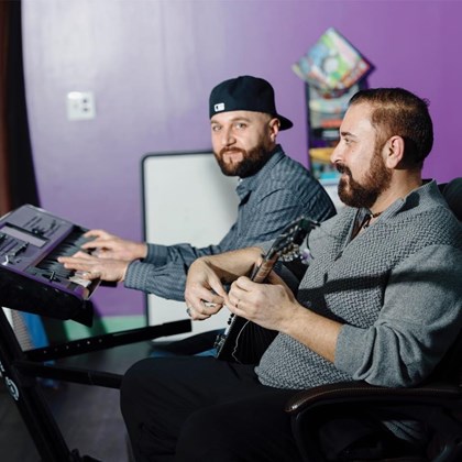 Two bearded young men play instruments while seated. The man in back plays keyboard and looks into the camera while the one in front plays a stringed instrument and looks to the left of the photo.