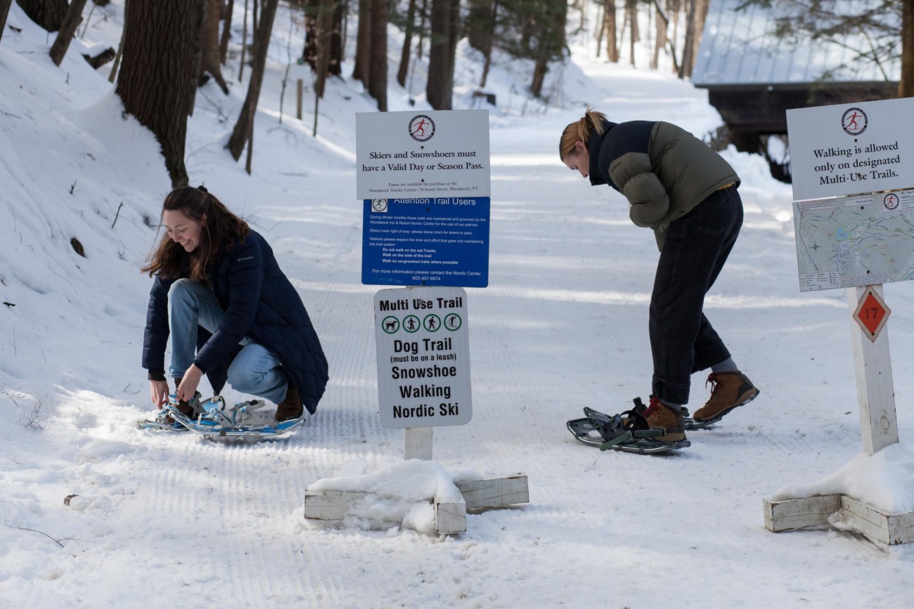 Two people put on snowshoes on groomed trail next to sign that has the following words legible: Multi-Use Trail, Dog Trail, Snowshoe, Walking, Nordic Ski,