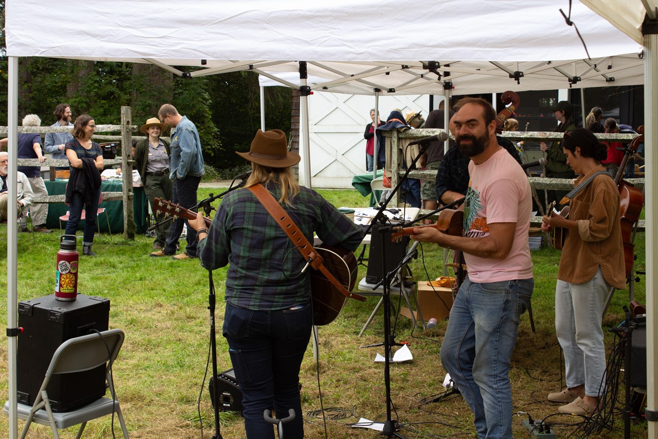 perspective of behind band performing under pop up tent, one band member looks at camera and the rest face towards audience