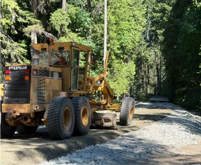 A grader works on a raised road bed in the forest.
