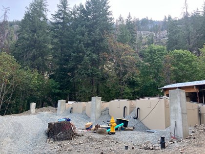Tan container structures, concrete posts, & fire hydrant amid spread gravel in front of a forest.