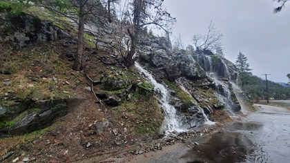 Water cascades off a rocky cliff face onto a road creating a standing puddle.