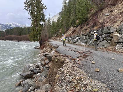 Two people in safety vests stand on a paved road that is falling into a lake with trees in backgroun
